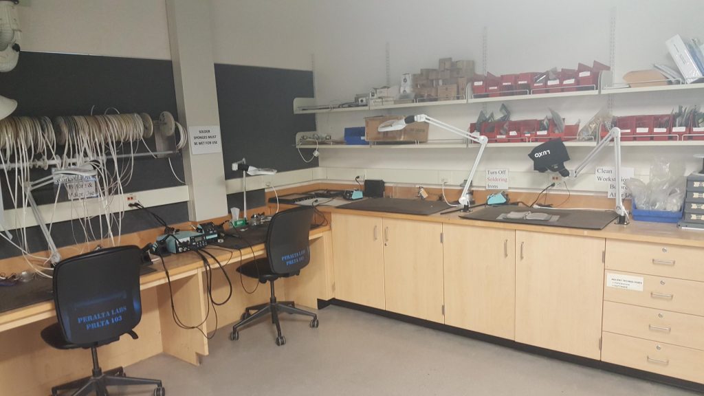 A workstation which includes two chairs a workbench and a rod with wires hanging from it. The space is furnished with a variety of racks, cupboards, and drawers for storing supplies.