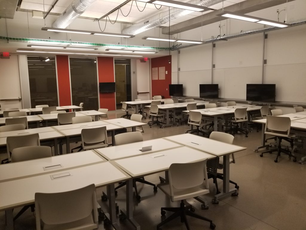 A classroom with several desks, chairs and TV screens.