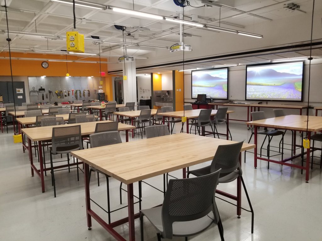 A classroom with desks, high rise chairs, projector screens and a pegboard with tools.
