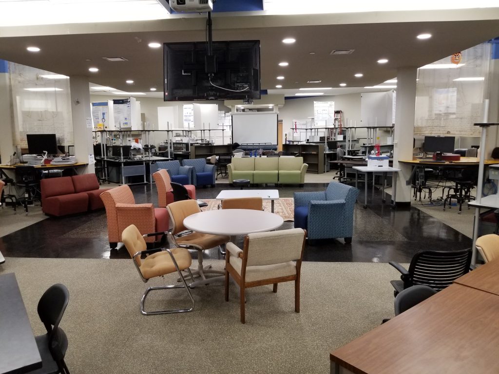 A seating area that consists of different colored sofas and chairs, a couple of monitors, desks and a white board.