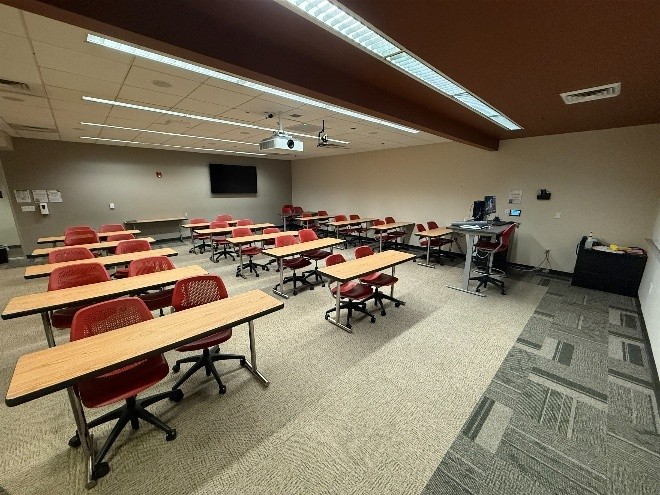 Picture from the northeast corner of the room showing six rows by three columns of desks - each with two Orange swivel chairs, and an automated stand-up desk as the podium in the Northwest corner of the room. 